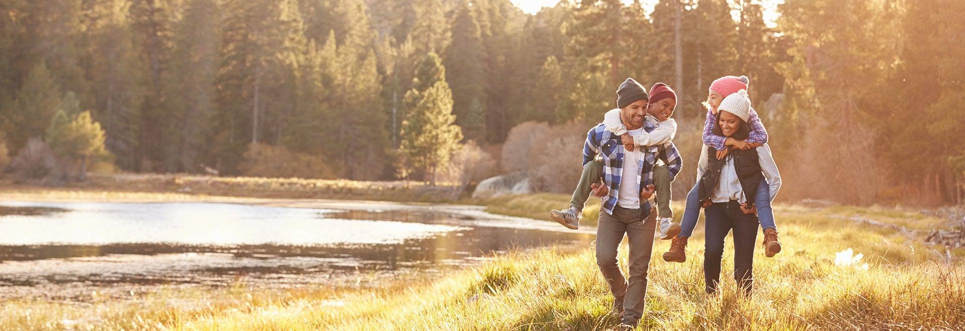 A family of four walks along a grassy lakeside path in a forested area. The parents carry their children on their backs, and everyone is dressed warmly in jackets and hats.