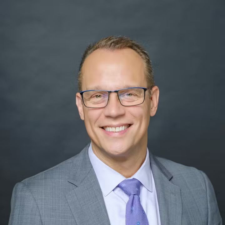 A smiling man with glasses wearing a suit and tie against a gray background.